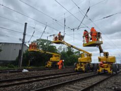 Network Rail to Deliver New Wixams Station Supporting Universal United Kingdom Resort Railway workers in orange high-visibility suits on yellow elevated platforms inspect overhead power lines above tracks under a cloudy sky.