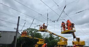 Network Rail to Deliver New Wixams Station Supporting Universal United Kingdom Resort Railway workers in orange high-visibility suits on yellow elevated platforms inspect overhead power lines above tracks under a cloudy sky.