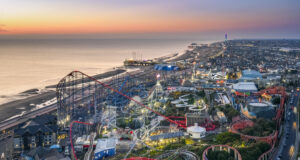 Pleasure Beach Resort wins eight at UK Theme Park Awards Aerial view of a seaside amusement park at sunset with roller coasters, a pier, and a city along the coastline.