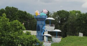 Paultons Park Pirate Ship & Windy Castle Ride Closed for Maintenance Colorful children's amusement ride with blue tower and cloud-shaped baskets, topped by pig characters seen above trees.