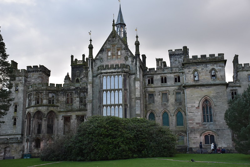 A medieval-style stone castle with tall towers and large arched windows, set behind a hedge on a grassy lawn