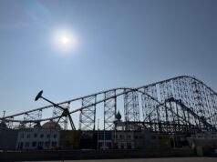 10 Things Every Family Should Know Before Visiting a Theme Park This Summer Bright sun over a clear blue sky with a large steel roller coaster towering above a row of colorful low-rise buildings beneath it.