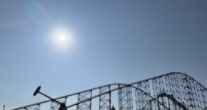 10 Things Every Family Should Know Before Visiting a Theme Park This Summer Bright sun over a clear blue sky with a large steel roller coaster towering above a row of colorful low-rise buildings beneath it.