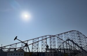 10 Things Every Family Should Know Before Visiting a Theme Park This Summer Bright sun over a clear blue sky with a large steel roller coaster towering above a row of colorful low-rise buildings beneath it.