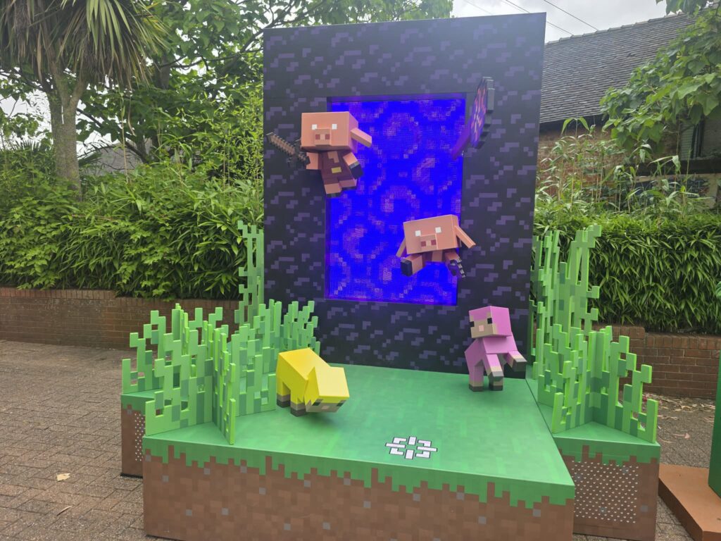 Minecraft-style display with pixelated green blocks, two pig characters, and a glowing purple portal in a garden setting