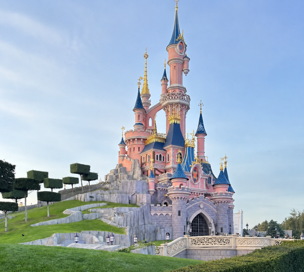 Pink fairy-tale castle with blue spires and gold trim atop a rocky hillside against a blue sky.