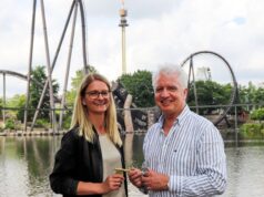 Heide Park Resort Welcomes New Managing Director as Peter Dunn Retires Smiling couple stands by a lake at an amusement park, with roller coasters in the background; the man holds a keyring.