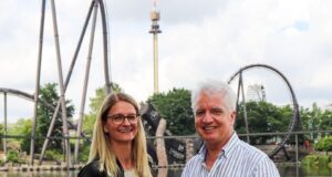 Heide Park Resort Welcomes New Managing Director as Peter Dunn Retires Smiling couple stands by a lake at an amusement park, with roller coasters in the background; the man holds a keyring.