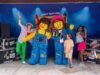 Everything You Need to Know About LEGO® Festival at LEGOLAND® Windsor 2026 People pose with two giant LEGO minifigures on a stage at a LEGO Festival, with a blue backdrop and stage lights.
