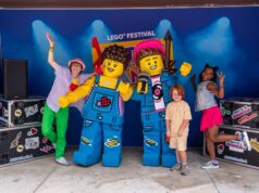 Everything You Need to Know About LEGO® Festival at LEGOLAND® Windsor 2026 People pose with two giant LEGO minifigures on a stage at a LEGO Festival, with a blue backdrop and stage lights.