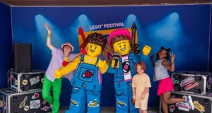 What’s Happening at UK Theme Parks This Week (Week of 20 July 2026) People pose with two giant LEGO minifigures on a stage at a LEGO Festival, with a blue backdrop and stage lights.