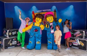 Everything You Need to Know About LEGO® Festival at LEGOLAND® Windsor 2026 People pose with two giant LEGO minifigures on a stage at a LEGO Festival, with a blue backdrop and stage lights.