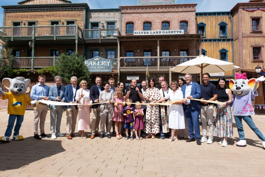 Group of adults and children pose with scissors for a ribbon-cutting in front of a rustic, storefront-lined western town set, with mascots on both sides.