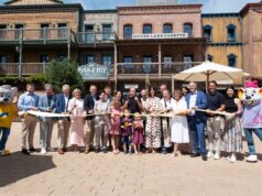 Europa-Park Opens Children’s House Little Heroes for Families Recovering from Cancer Group of adults and children pose with scissors for a ribbon-cutting in front of a rustic, storefront-lined western town set, with mascots on both sides.