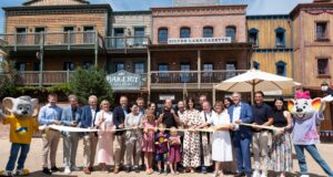 Europa-Park Opens Children’s House Little Heroes for Families Recovering from Cancer Group of adults and children pose with scissors for a ribbon-cutting in front of a rustic, storefront-lined western town set, with mascots on both sides.