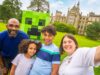 What’s Happening at UK Theme Parks This Weekend (18–19 July 2026) Family of four smiling for a selfie on a sunny lawn, with a life-size Minecraft Creeper head behind them and a castle-like building in the background.