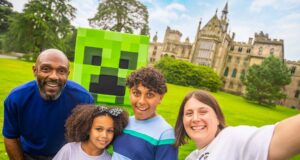 What’s Happening at UK Theme Parks This Weekend (18–19 July 2026) Family of four smiling for a selfie on a sunny lawn, with a life-size Minecraft Creeper head behind them and a castle-like building in the background.