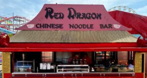 Brand-New Red Dragon Chinese Noodle Bar Opens at Barry Island Pleasure Park Bright red noodle bar stall with a large sign reading 'Red Dragon Chinese Noodle Bar' and workers behind the counter in an amusement park setting.