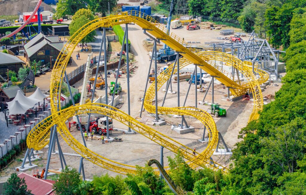 Aerial view of a yellow roller coaster under construction with scaffolding, dirt ground, and construction vehicles nearby.