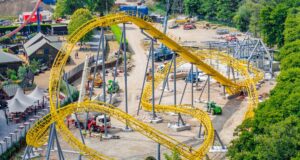 Serpent’s Curse Construction Update: More Track Installed at Paultons Park Aerial view of a yellow roller coaster under construction with scaffolding, dirt ground, and construction vehicles nearby.