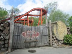 The Classic Log Flumes You Can Still Ride in the UK This Summer Wooden gate in front of an orange roller coaster track rising behind, with stacked logs on the left and a carved tree stump on the right.