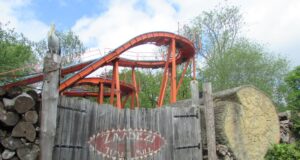 The Classic Log Flumes You Can Still Ride in the UK This Summer Wooden gate in front of an orange roller coaster track rising behind, with stacked logs on the left and a carved tree stump on the right.