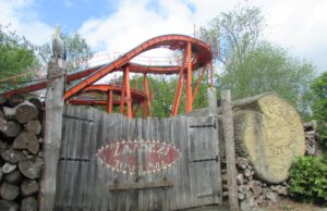 The Classic Log Flumes You Can Still Ride in the UK This Summer Wooden gate in front of an orange roller coaster track rising behind, with stacked logs on the left and a carved tree stump on the right.