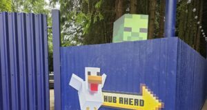 What’s Happening at UK Theme Parks This Weekend (25–26 July 2026) Blue corrugated fence with a Minecraft-style pixel art sign showing a white blocky figure and the yellow 'HUB AHEAD' text.