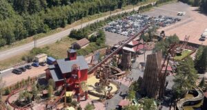 Fireline Redwood Officially Opens at Fraispertuis-City for the Park’s 60th Anniversary Aerial view of a rustic amusement park with red wooden buildings, roller coasters, and a large parking lot nearby.