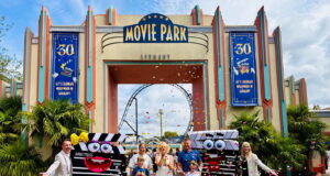 Movie Park Germany Welcomes its 40-millionth Guest Group of people posing with two cartoon clapperboard mascots in front of Movie Park Germany entrance as confetti falls and a roller coaster is visible in the background.