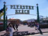 Great Yarmouth Pleasure Beach Offers £10 Thursday Wristbands Entrance to Pleasure Beach with a large decorative sign; people walk along a sunny boardwalk with stalls and a carousel in the background.
