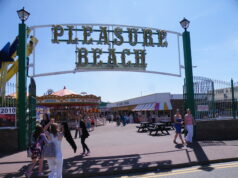 Great Yarmouth Pleasure Beach Offers ยฃ10 Thursday Wristbands Entrance to Pleasure Beach with a large decorative sign; people walk along a sunny boardwalk with stalls and a carousel in the background.