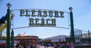 Great Yarmouth Pleasure Beach Offers £10 Thursday Wristbands Entrance to Pleasure Beach with a large decorative sign; people walk along a sunny boardwalk with stalls and a carousel in the background.