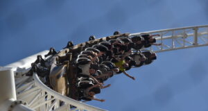 Guests Remember Experiences — Not Ride Specs Riders on a white roller coaster car mid-loop against a bright blue sky, arms outstretched.