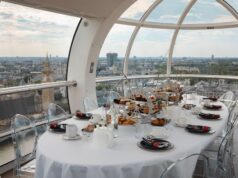 EXPIRED: Savour the Skyline: Get 15% Off London Eye Afternoon Tea Luxurious round dining table set with pastries inside a glass capsule overlooking a city skyline with iconic towers, clear chairs and white tableware