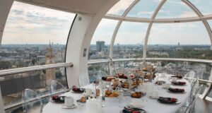 Savour the Skyline: Get 15% Off London Eye Afternoon Tea Luxurious round dining table set with pastries inside a glass capsule overlooking a city skyline with iconic towers, clear chairs and white tableware