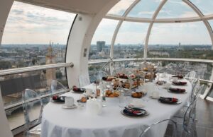 Savour the Skyline: Get 15% Off London Eye Afternoon Tea Luxurious round dining table set with pastries inside a glass capsule overlooking a city skyline with iconic towers, clear chairs and white tableware