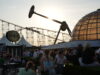 Pleasure Beach Resort Hands Out 1,000 Free Eclipse Glasses to Visitors People gather at an outdoor amusement park at sunset, with roller coaster tracks and a glass-domed building in the background and booths nearby.