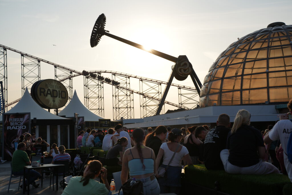 People gather at an outdoor amusement park at sunset, with roller coaster tracks and a glass-domed building in the background and booths nearby.