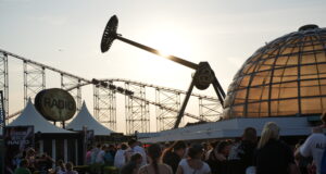 Pleasure Beach Resort Hands Out 1,000 Free Eclipse Glasses to Visitors People gather at an outdoor amusement park at sunset, with roller coaster tracks and a glass-domed building in the background and booths nearby.