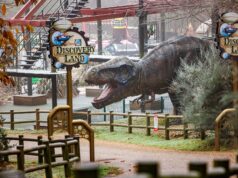 10 Things You Didn’t Know About ROARR! Dinosaur Adventure Huge dinosaur statue in a theme park area with signage reading 'Discovery Land' and a red ride track overhead.