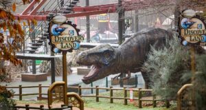 10 Things You Didn’t Know About ROARR! Dinosaur Adventure Huge dinosaur statue in a theme park area with signage reading 'Discovery Land' and a red ride track overhead.