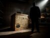 Final Exhibit Behind the Scenes: Go Behind the Scenes of Scarefest’s Newest Maze Silhouetted man stands in a dark warehouse beside a large wooden crate, spotlight shining from above.