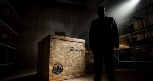 Final Exhibit Behind the Scenes: Go Behind the Scenes of Scarefest’s Newest Maze Silhouetted man stands in a dark warehouse beside a large wooden crate, spotlight shining from above.