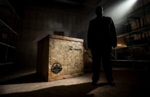 Final Exhibit Behind the Scenes: Go Behind the Scenes of Scarefest’s Newest Maze Silhouetted man stands in a dark warehouse beside a large wooden crate, spotlight shining from above.