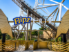 Flipz Takes Over Hyperia at Thorpe Park for Three Weeks Entrance sign for the Flipz area of an amusement park with gold gate panels and a roller coaster track in the background, bright blue sky