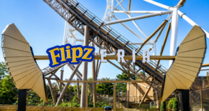 Flipz Takes Over Hyperia at Thorpe Park for Three Weeks Entrance sign for the Flipz area of an amusement park with gold gate panels and a roller coaster track in the background, bright blue sky