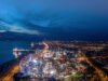Pleasure Beach Resort to Host Mass Gathering for Upcoming Eclipse Aerial night view of a coastal city with a brightly lit amusement park and ferris wheel along the waterfront."