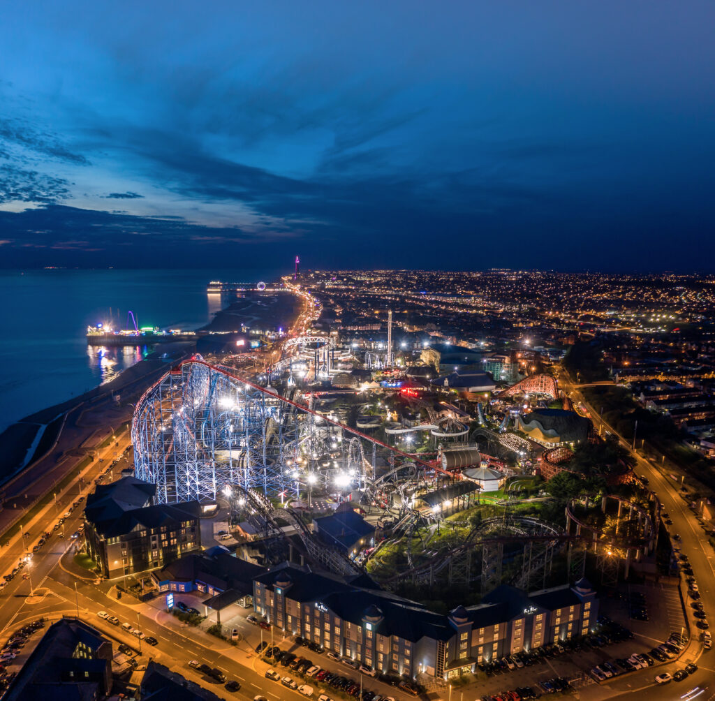 Aerial night view of a coastal city with a brightly lit amusement park and ferris wheel along the waterfront."