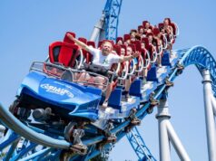 Rob Beckett discovers Europa-Park – Germany’s Largest Theme Park People riding a blue roller coaster with red seats, arms raised, against a clear blue sky, smiling and laughing as they zoom along the track.
